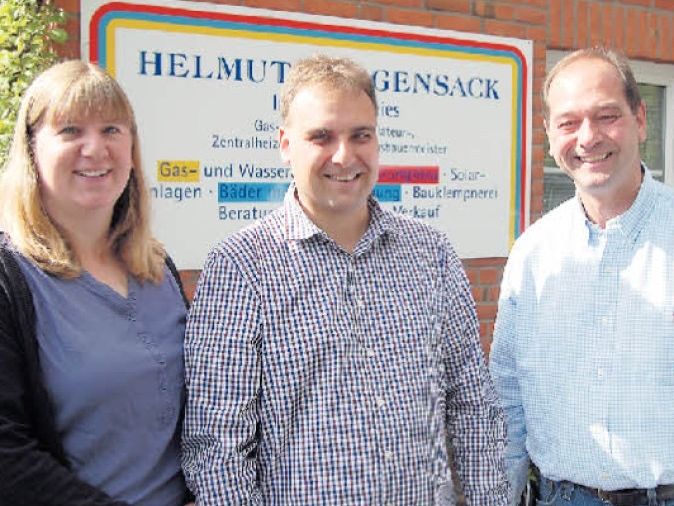 Drei Personen stehen vor einem Firmenschild von Helmut Gensack, einem Unternehmen für Gas-, Wasser- und Heizungsanlagen.
