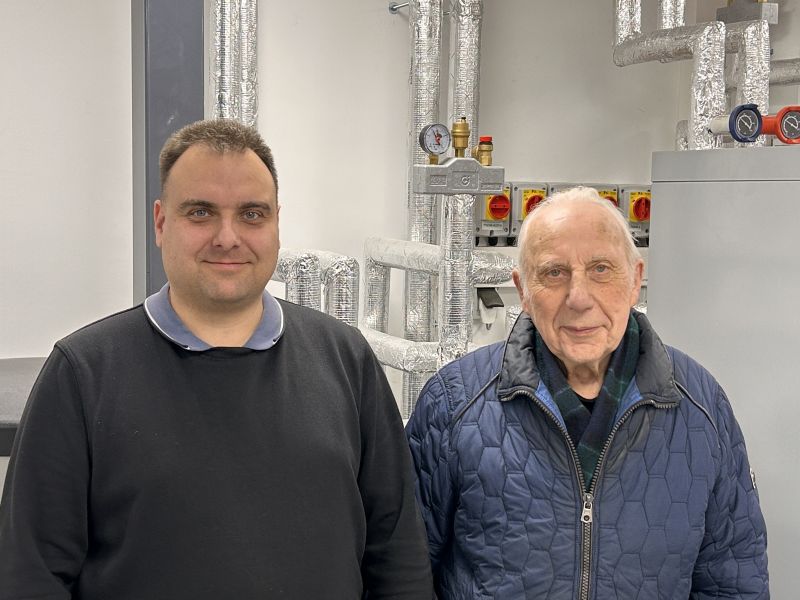 Zwei Männer stehen vor einer Wand mit isolierten Rohren und Messgeräten in einem technischen Raum