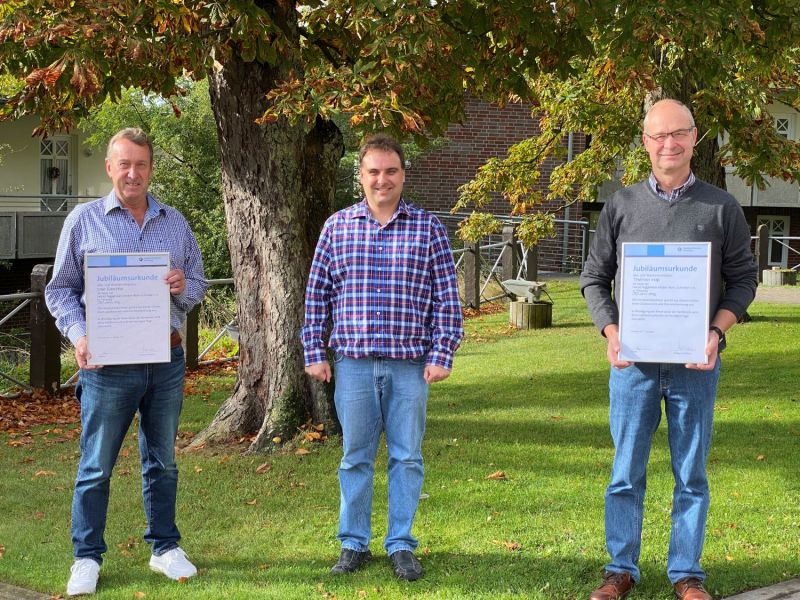 Drei Männer stehen auf einer Wiese vor einem Baum, zwei halten Urkunden mit der Überschrift 'Jubiläumsurkunde'